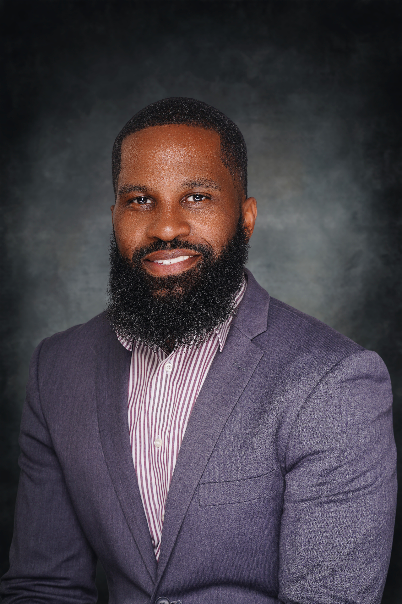 Professional studio headshot of a man in a blazer against a dark textured backdrop